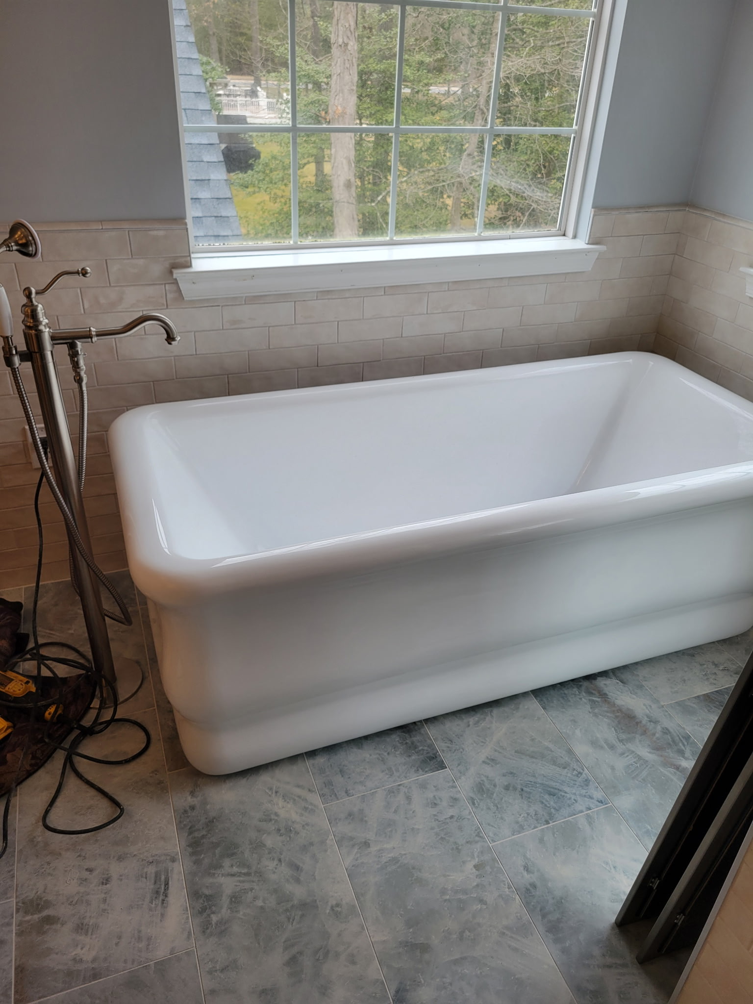 Freestanding white bathtub beneath a large window with a blue-gray tiled floor