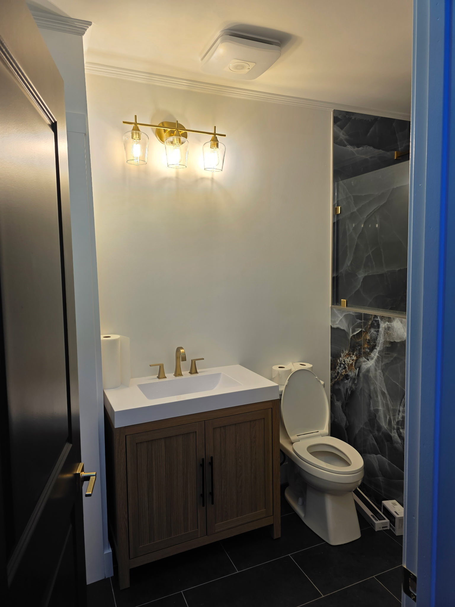Finished bathroom with light wood vanity, dark stone-look shower tile, and gold fixtures