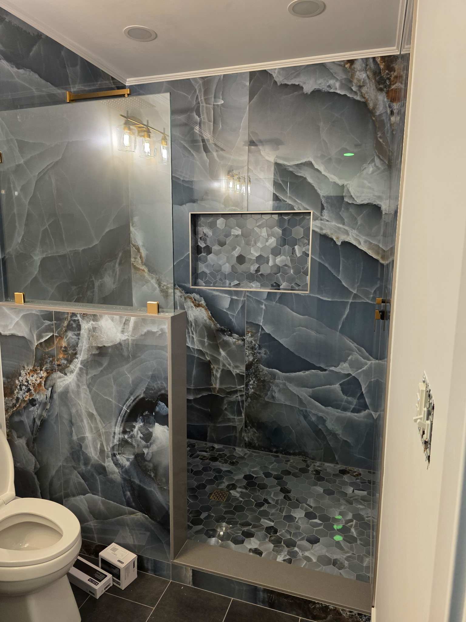 Custom walk-in shower with dramatic stone-look tile, hexagon niche, glass panel, and gold accents